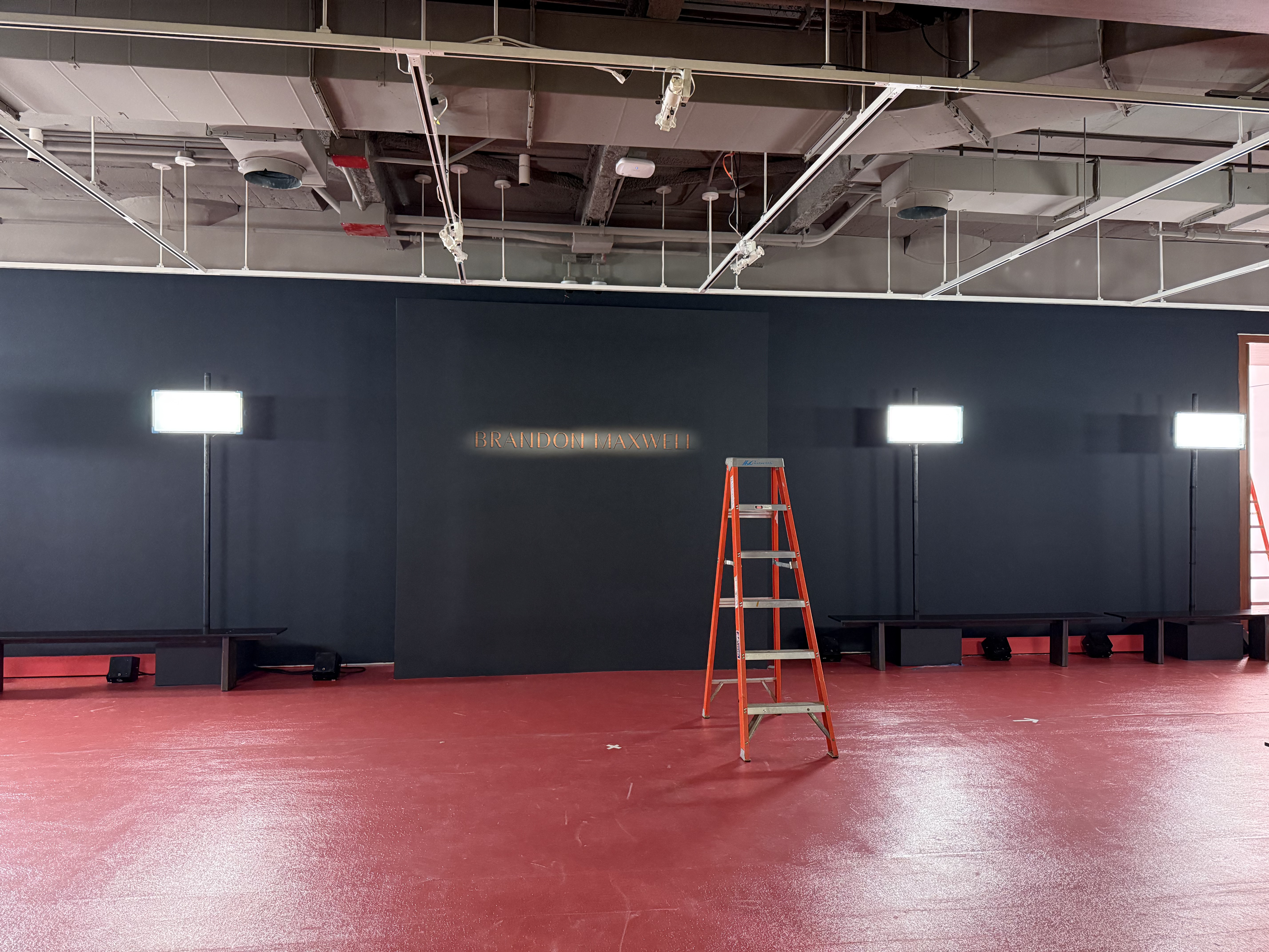 Backstage step-and-repeat wall with the Brandon Maxwell name lit up, a ladder set up for install.
