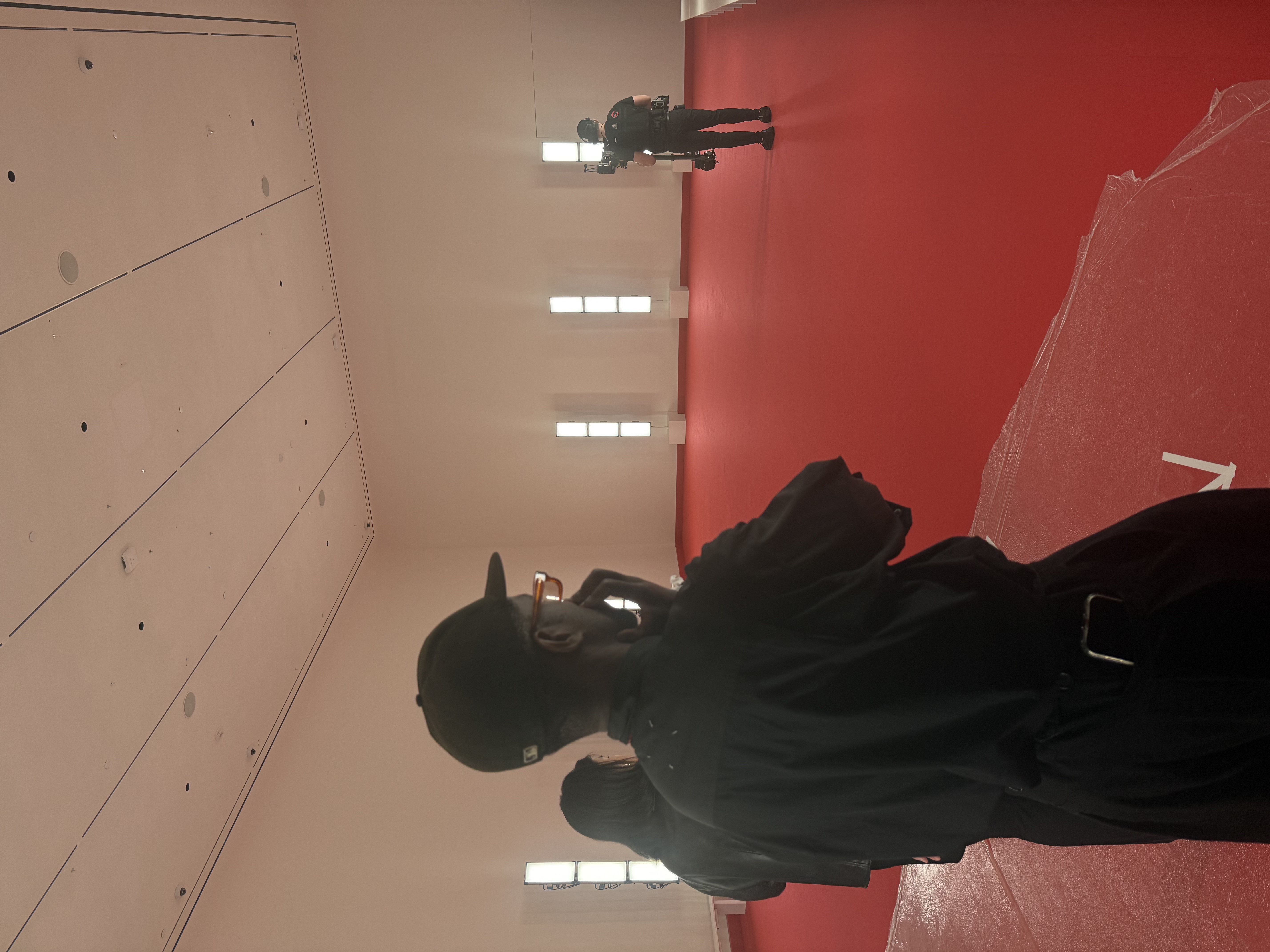 Backstage production crew filming against a red wall, with plastic-wrapped furniture nearby.