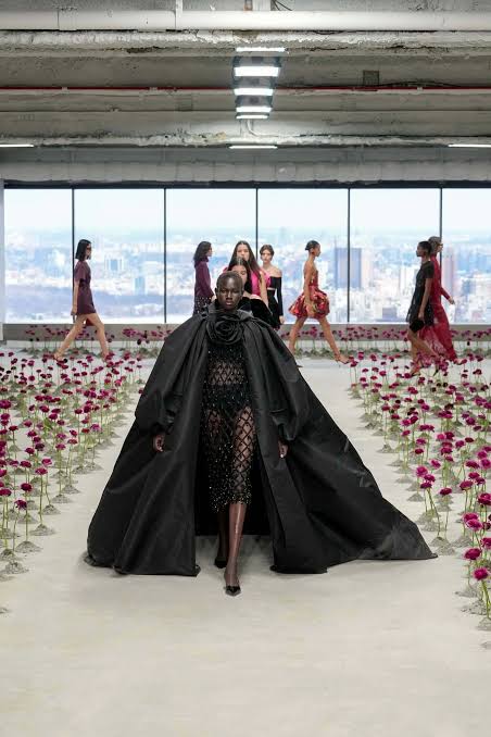 Model in a voluminous black gown leading a group down the runway through the flower field, city skyline behind.
