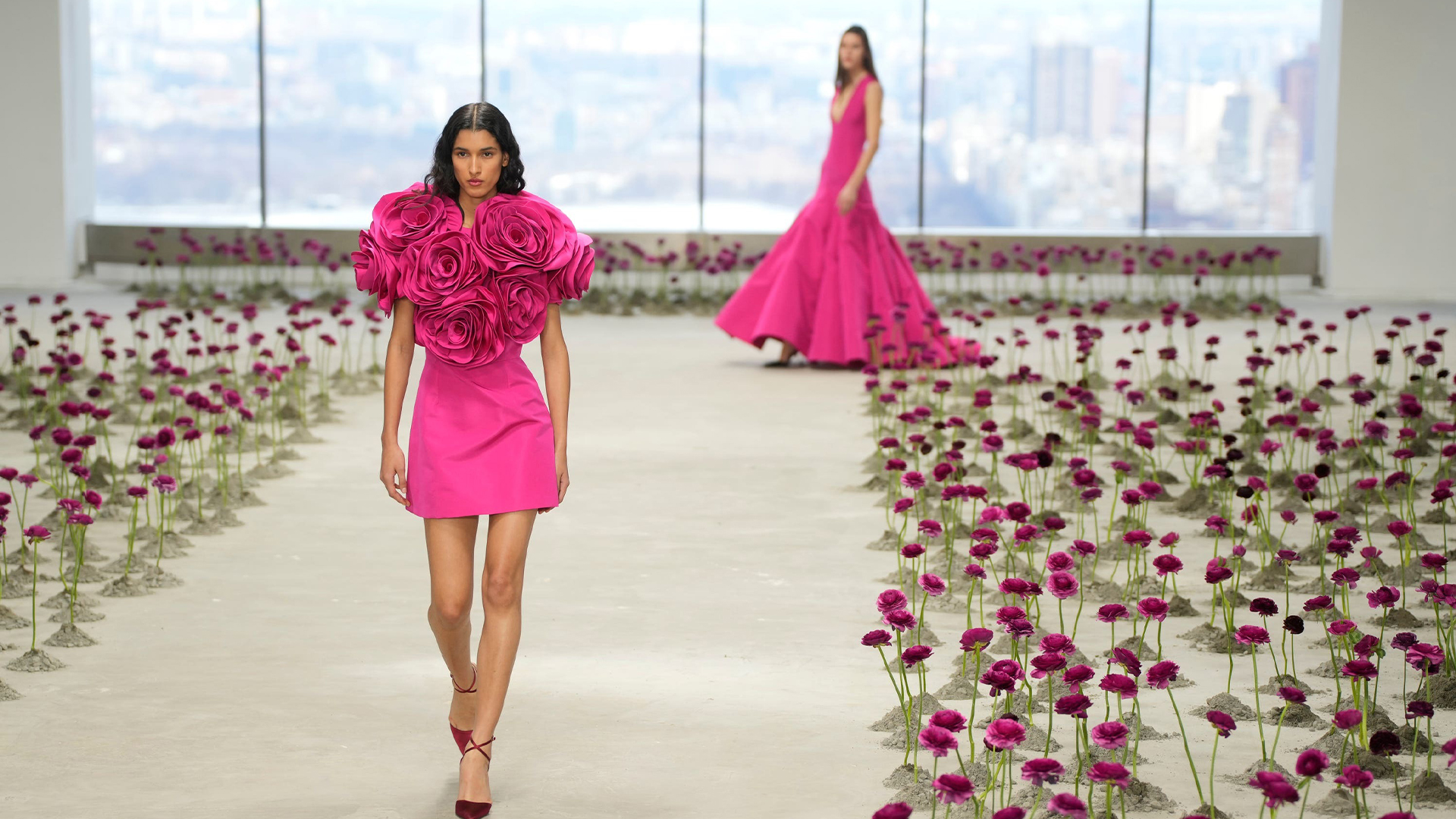 Model in a hot pink mini dress with a 3D rose bodice walking the runway, another model in a pink gown behind her.