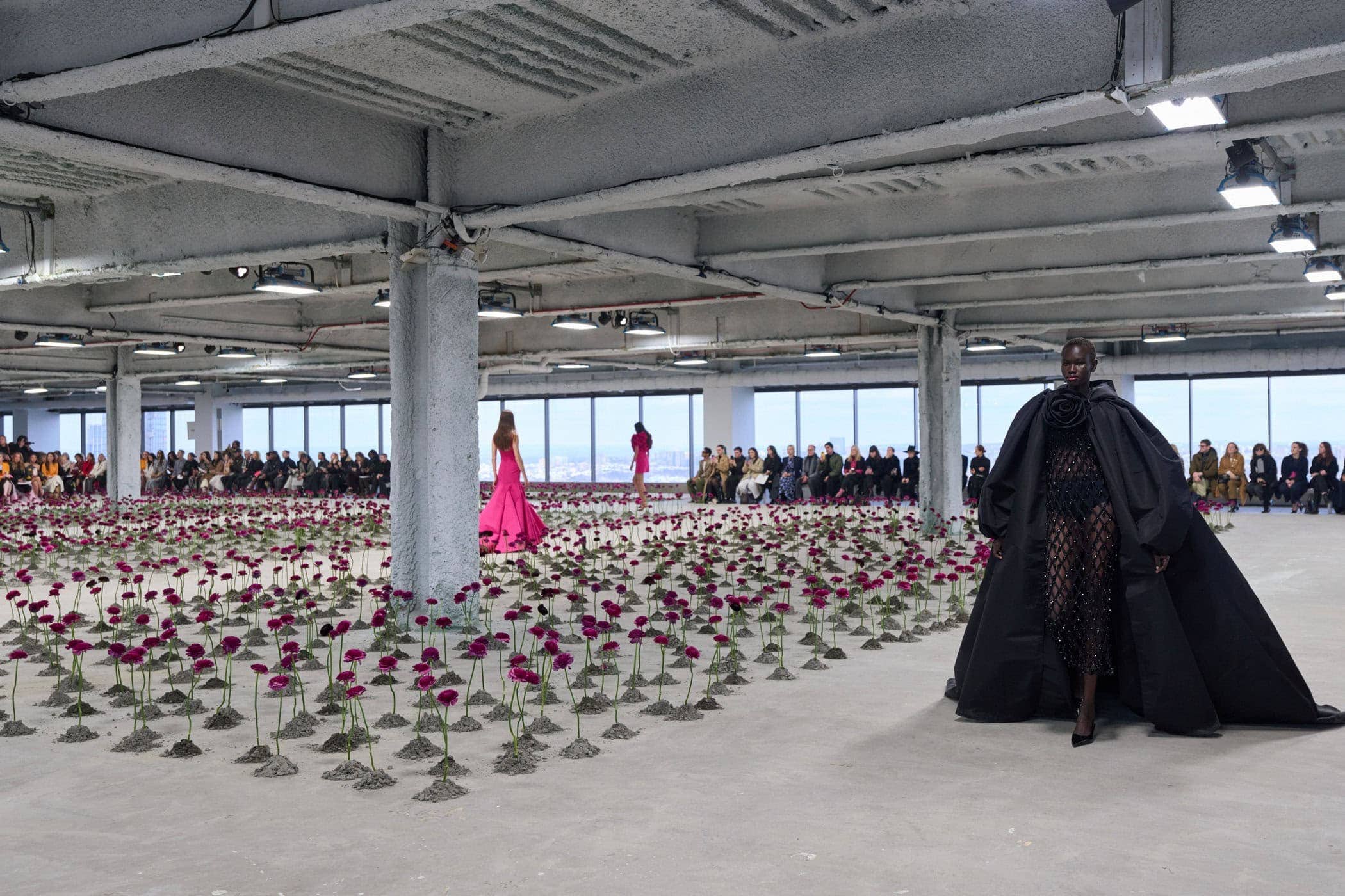 Model in a voluminous black gown with a rose-appliqued shoulder walking the runway through a field of magenta ranunculus planted in the concrete floor, guests seated on either side.