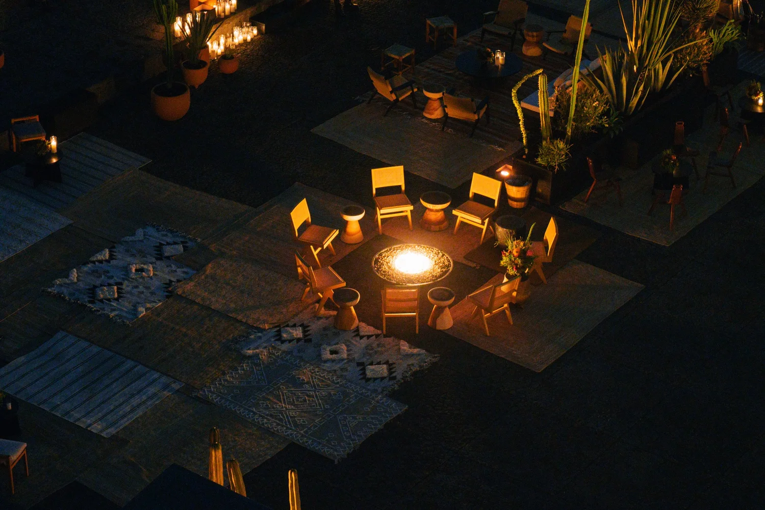 Aerial view of a candlelit fire-pit lounge, chairs and layered rugs arranged in a circle on the museum plaza.