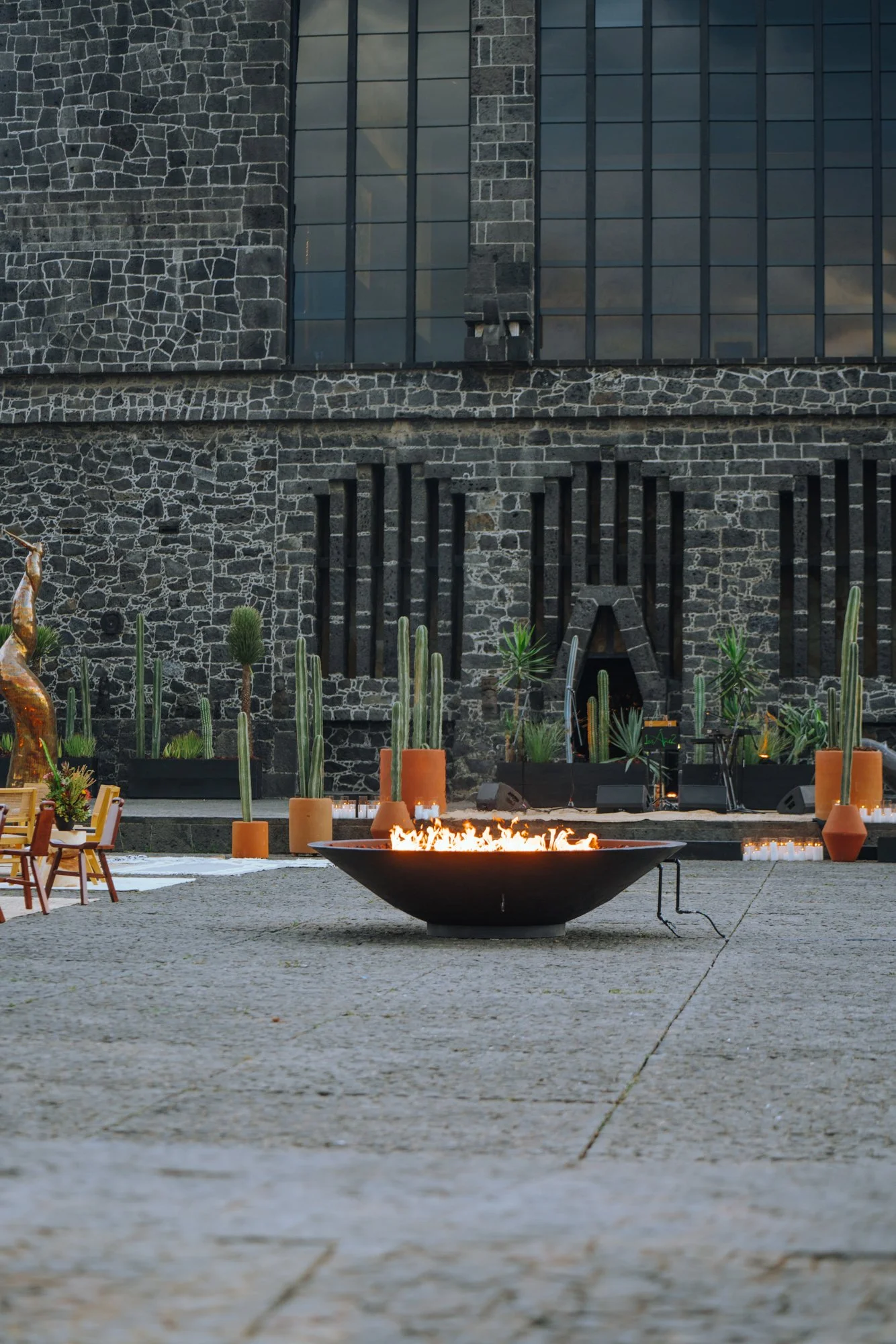 A fire bowl burning in front of the museum's stone facade, with cactus planters and a bronze sculpture nearby.