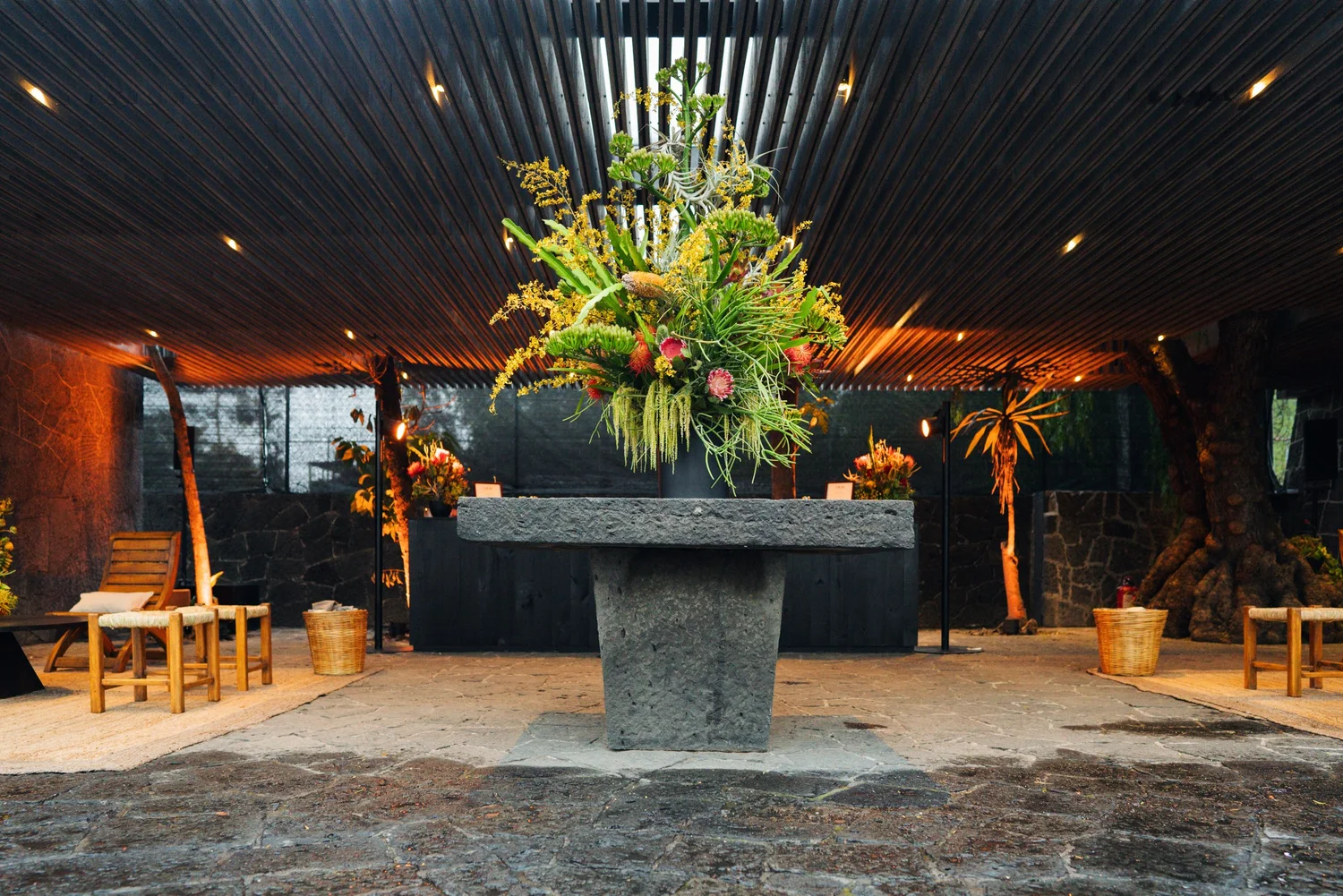 A tall floral installation on a carved stone table beneath a slatted wood canopy.