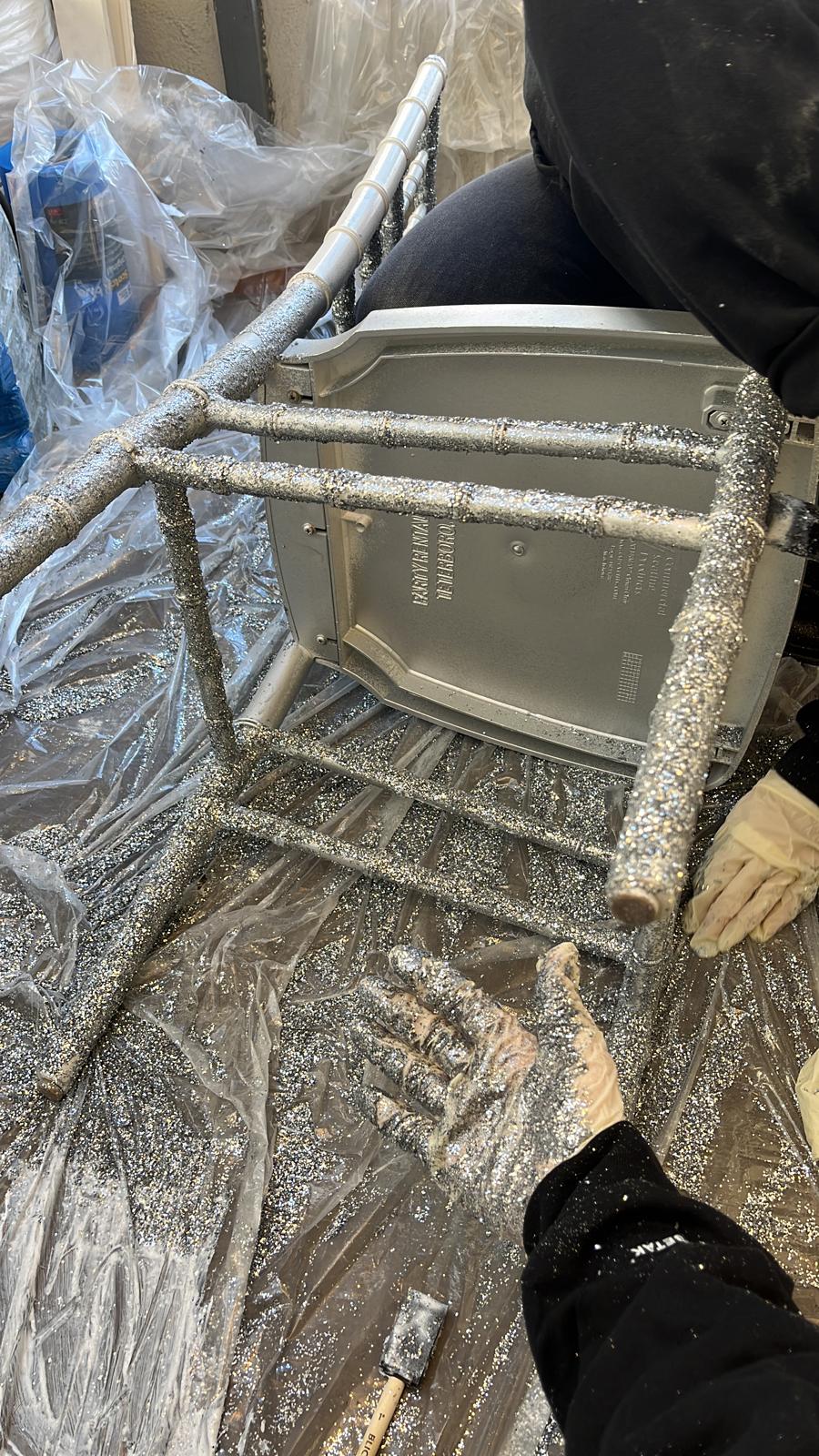 Hand applying silver glitter to a chair frame, wrapped in plastic sheeting during prep.