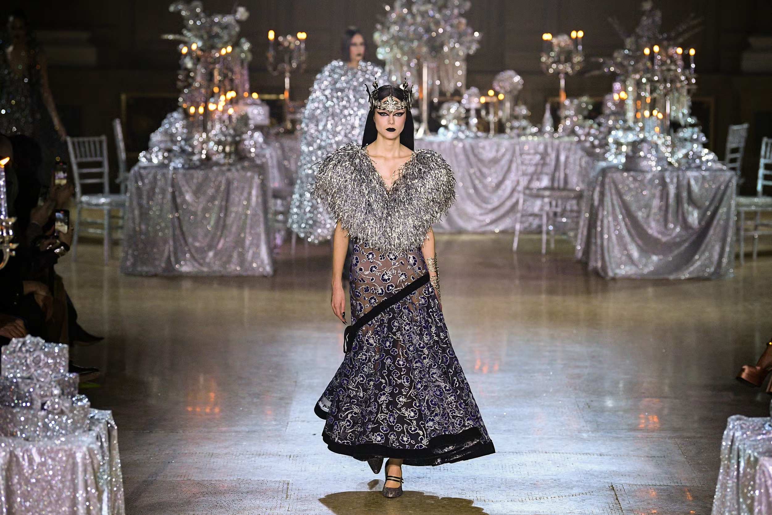 Model in an embroidered gown with feathered silver shoulders and a crown headpiece, walking past a glittered tablescape at the Rodarte Fall/Winter 23 show.