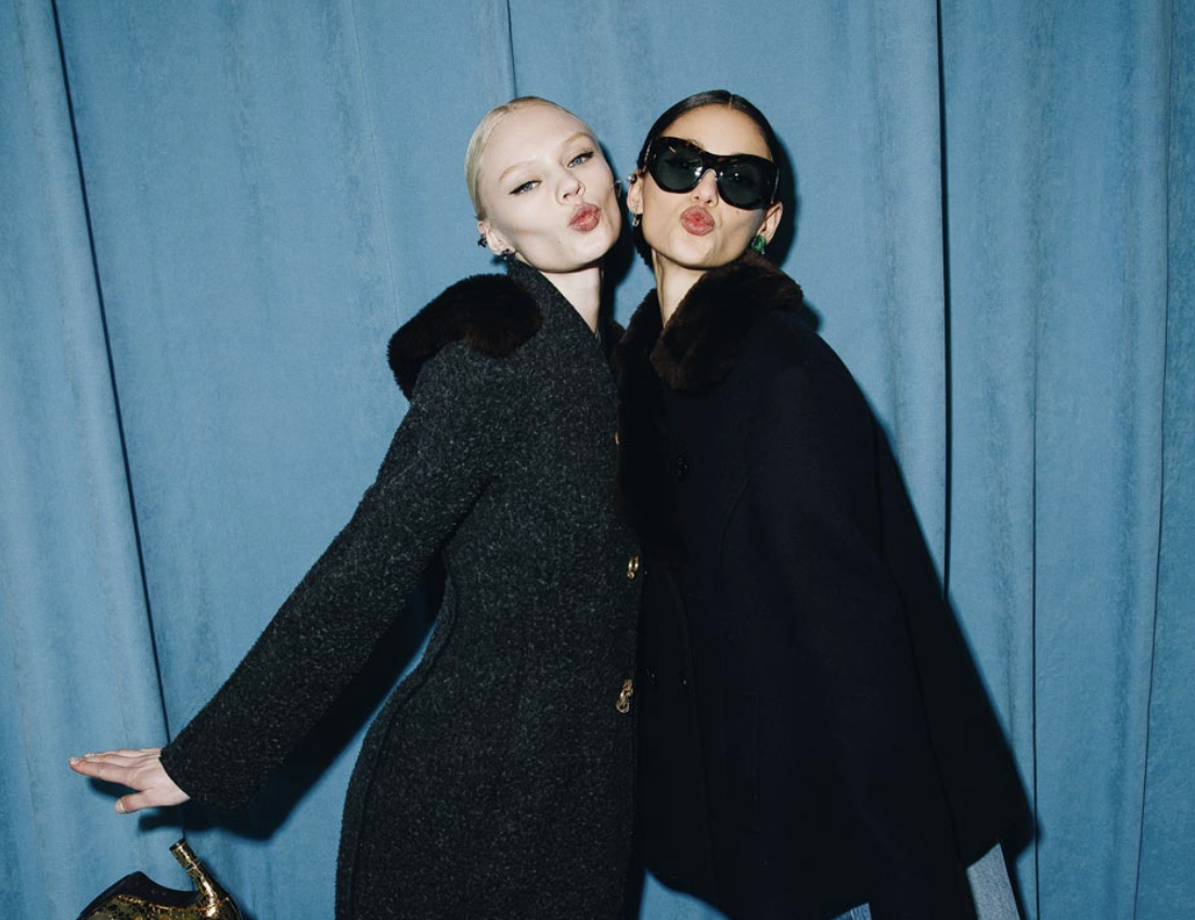 Two models backstage in coats with fur-trimmed collars, posing against a blue curtain.