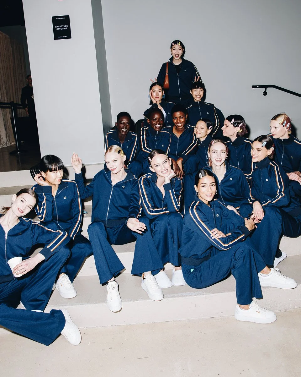 Models backstage in matching navy tracksuits, seated together on a staircase.