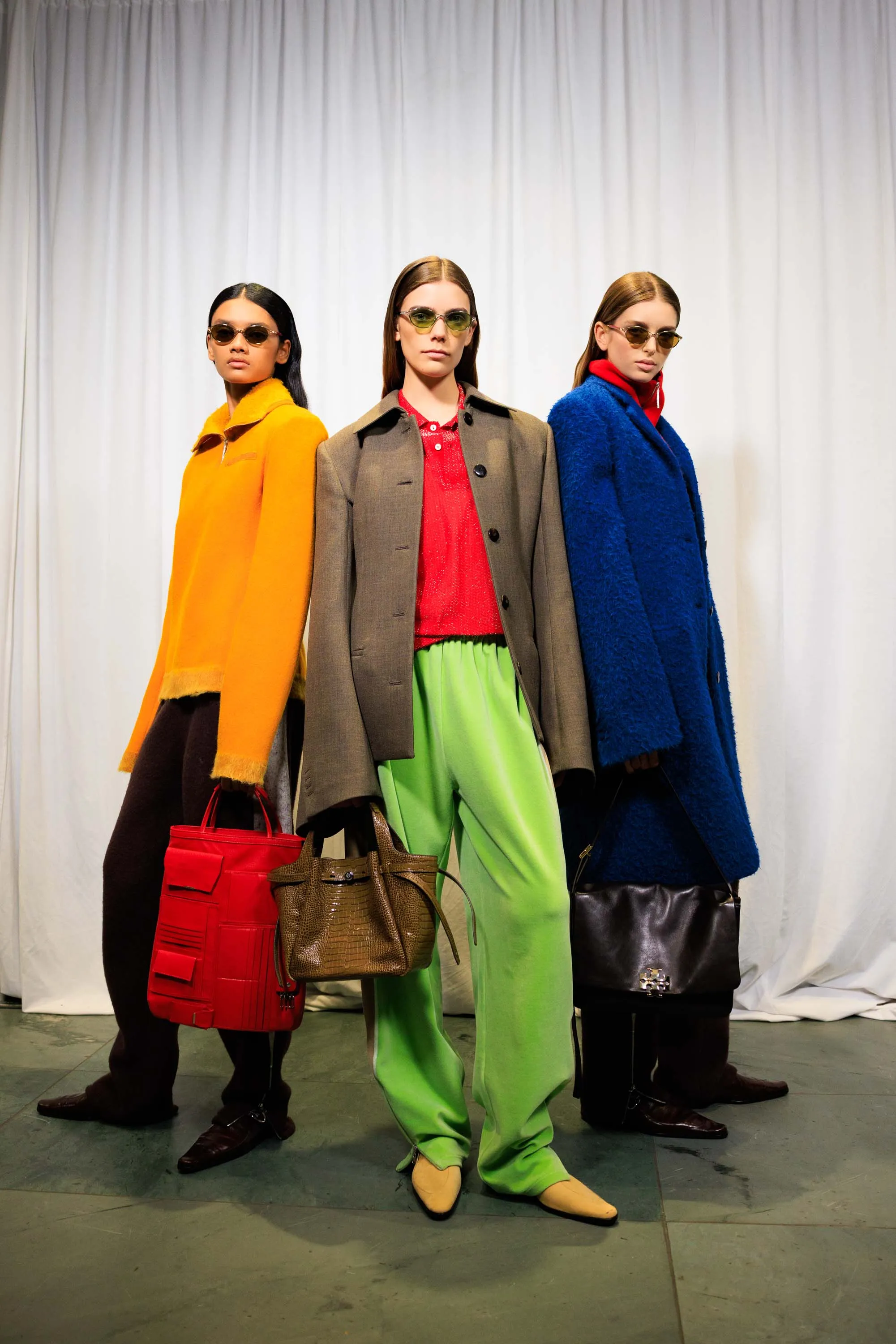 Three models in bold colorblocked coats and trousers — orange, grey, and blue — each carrying a structured handbag.