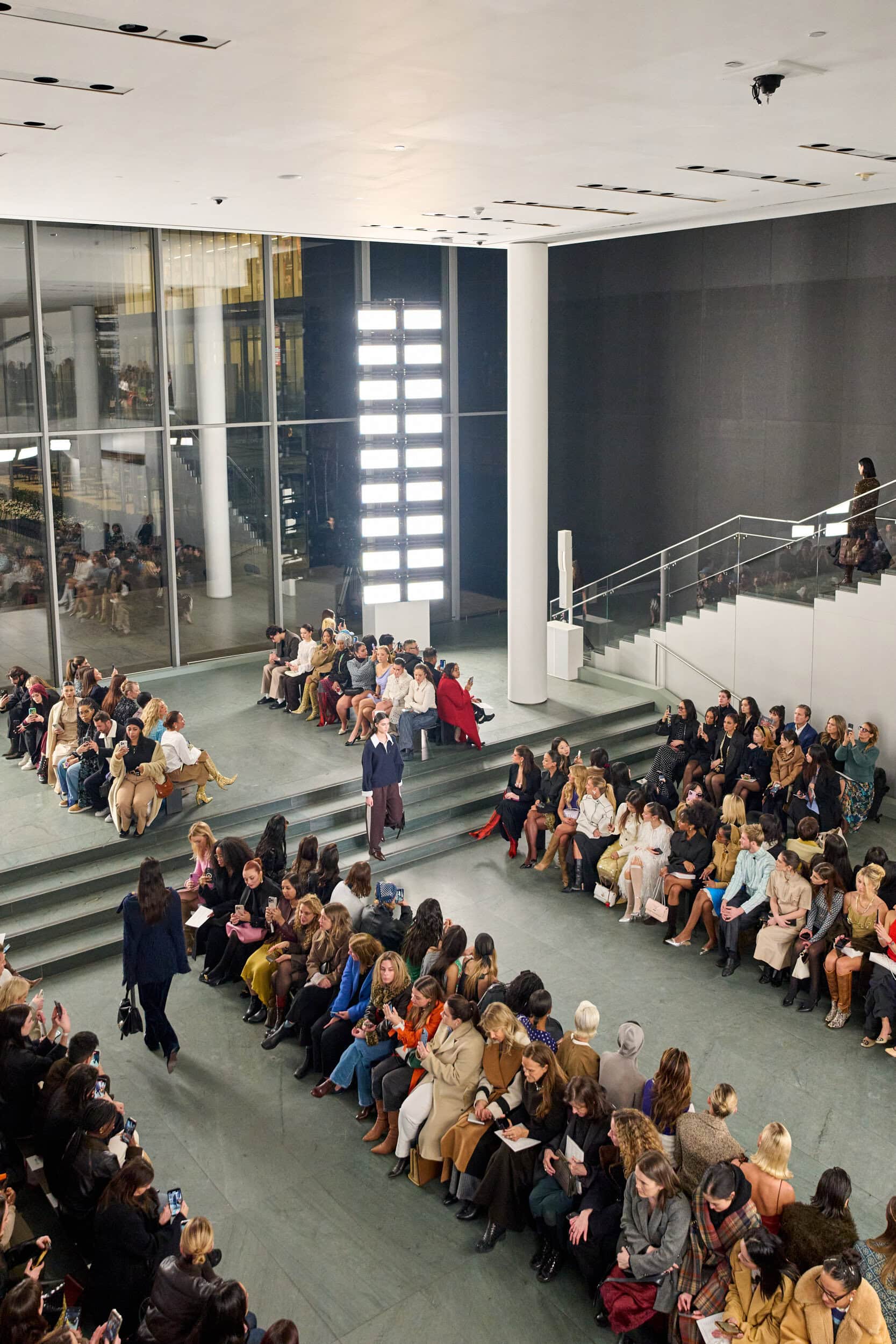 Overhead view of the MoMA atrium runway with a model descending the staircase past seated guests.