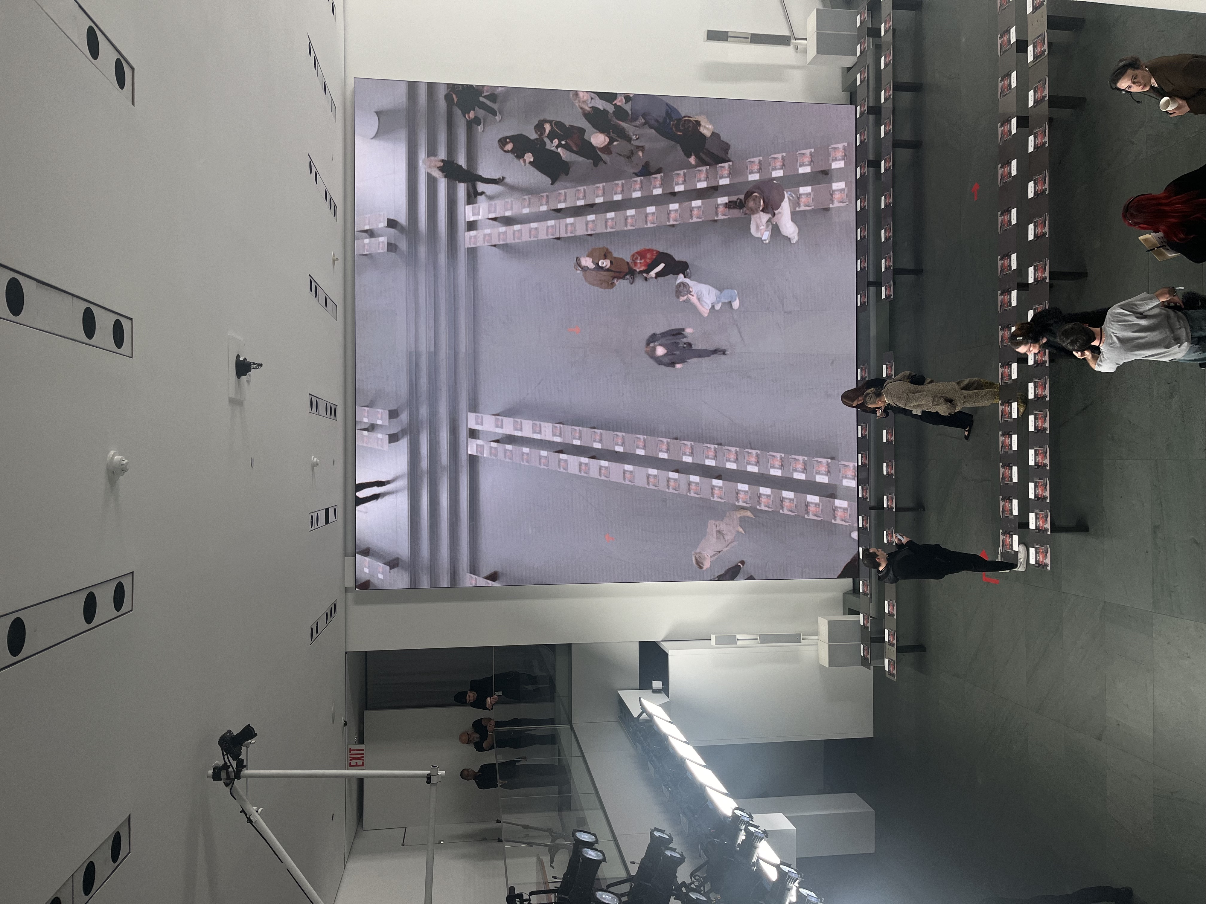 Overhead camera feed on a screen showing guests arriving over the seat-card-lined runway, lighting rig in view.