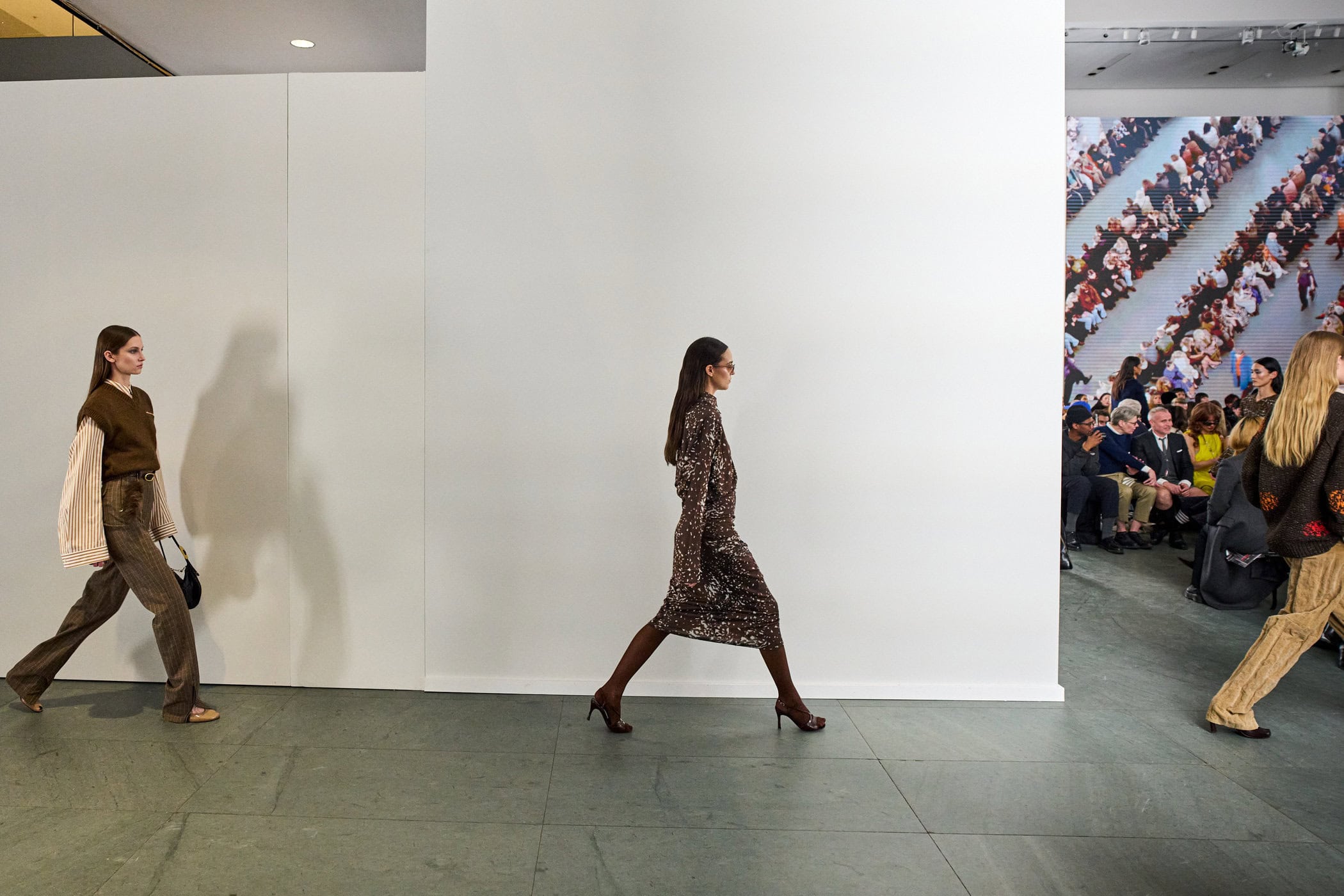Models walking past a white dividing wall with a live video feed of the seated audience, at the Tory Burch Fall/Winter 25 show.