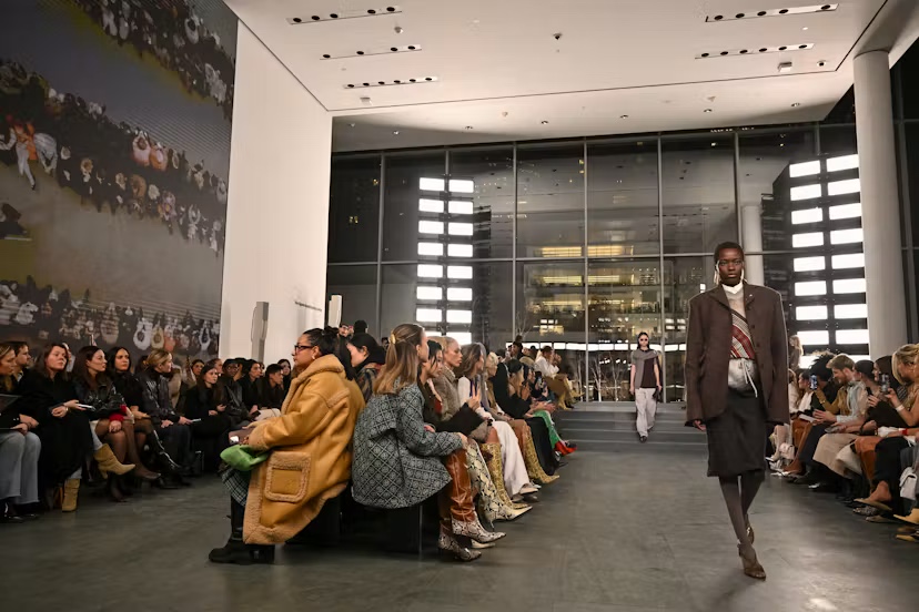 Model in a tailored brown coat walking the runway in MoMA's glass-walled lobby atrium, seated guests on either side.